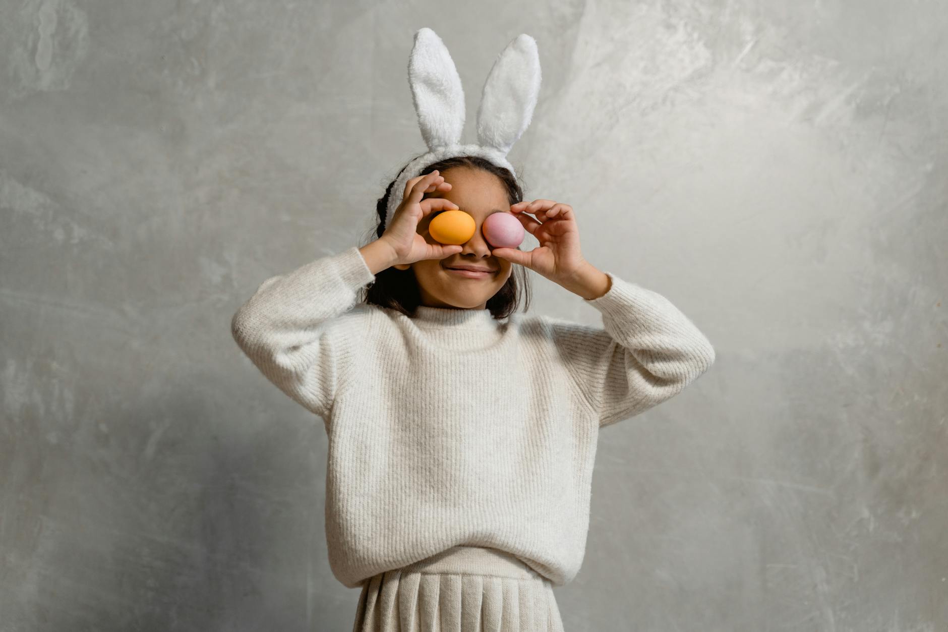 girl wearing easter bunny headwear covering her eyes with easter eggs